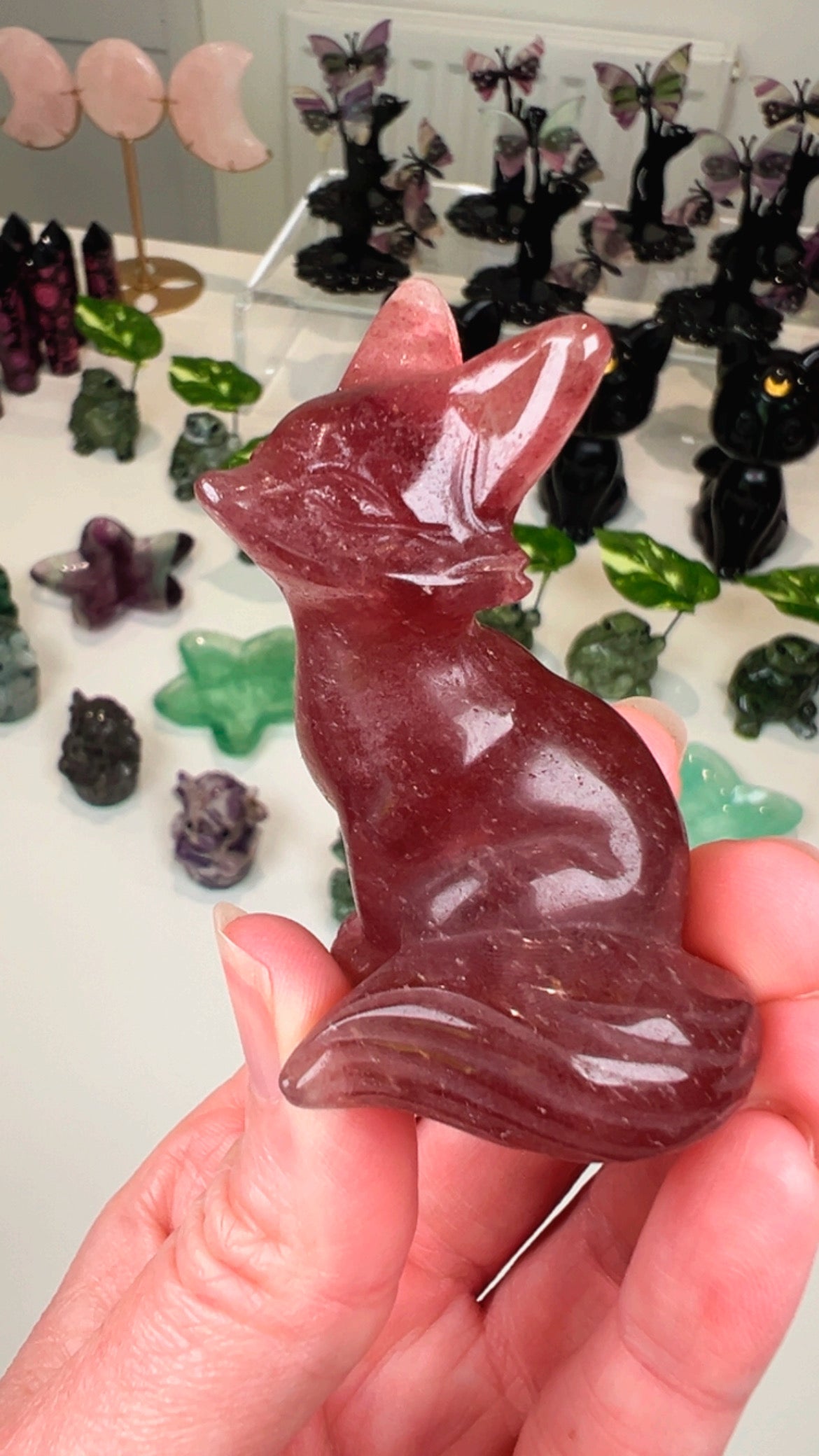 A hand holding a polished reddish-brown crystal fox figurine, with other colorful crystal formations displayed in the blurred background.