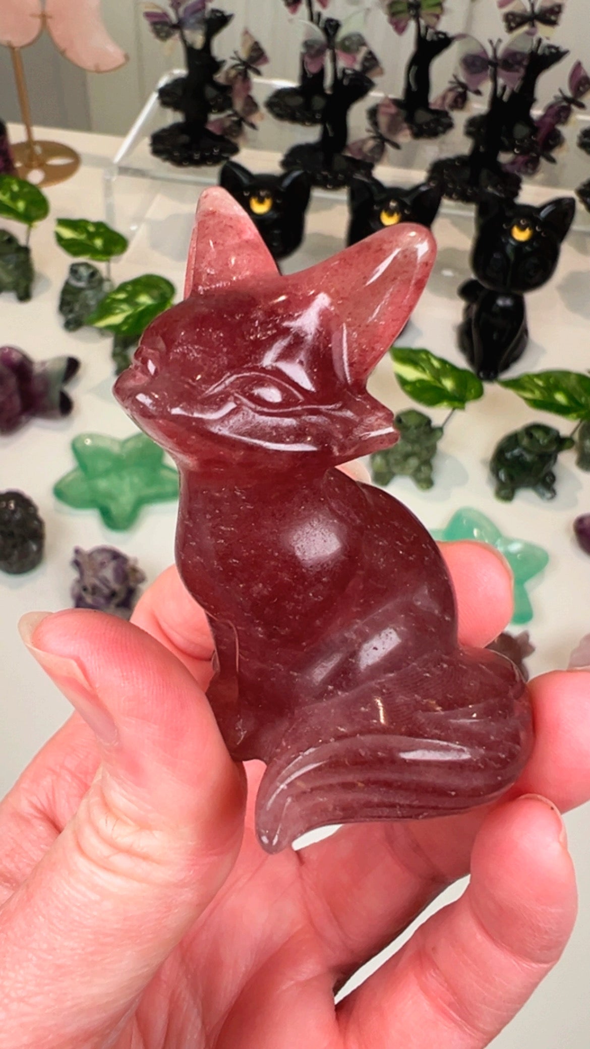 A hand holds a polished pinkish-red crystal fox figurine, with other colorful crystal carvings displayed in the blurred background.