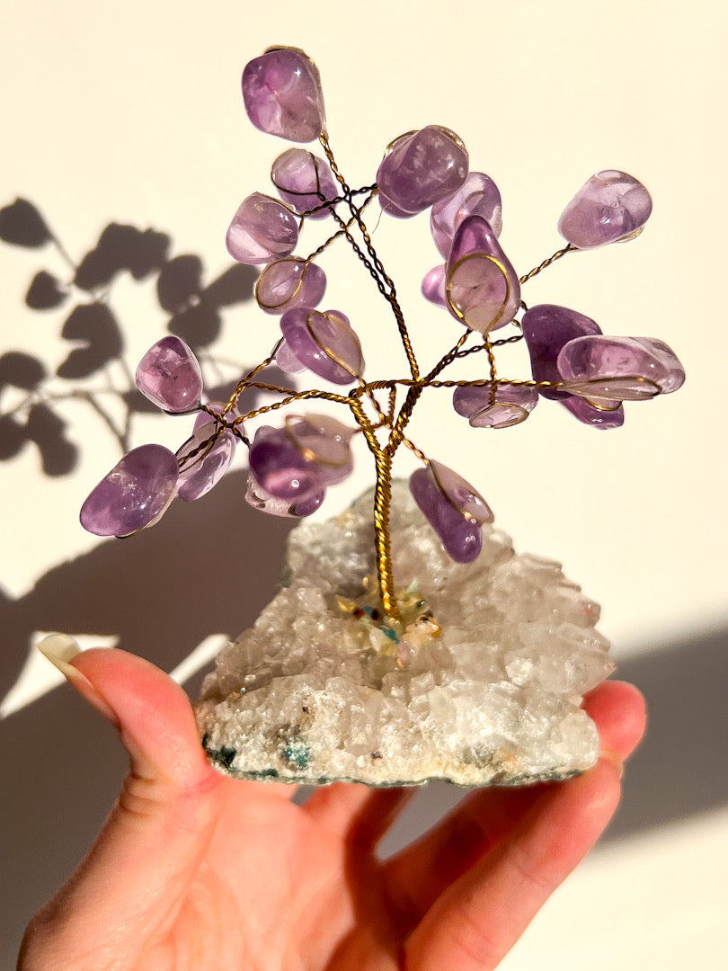 Hand holding a crystal tree with purple stones on a clear crystal base against a neutral background