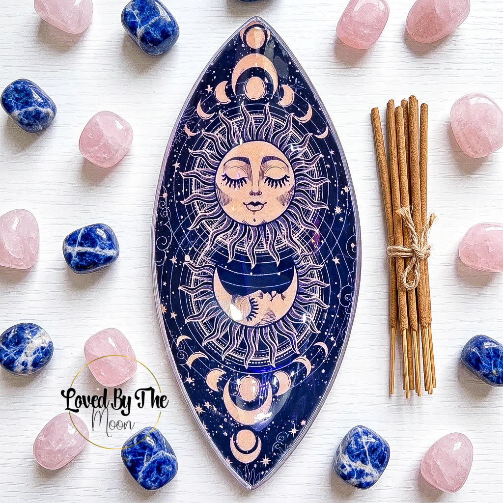 An oval moon-phase incense stick holder with a celestial sun and moon design, styled on a white wood background with pink and blue crystals and a bundle of incense sticks.