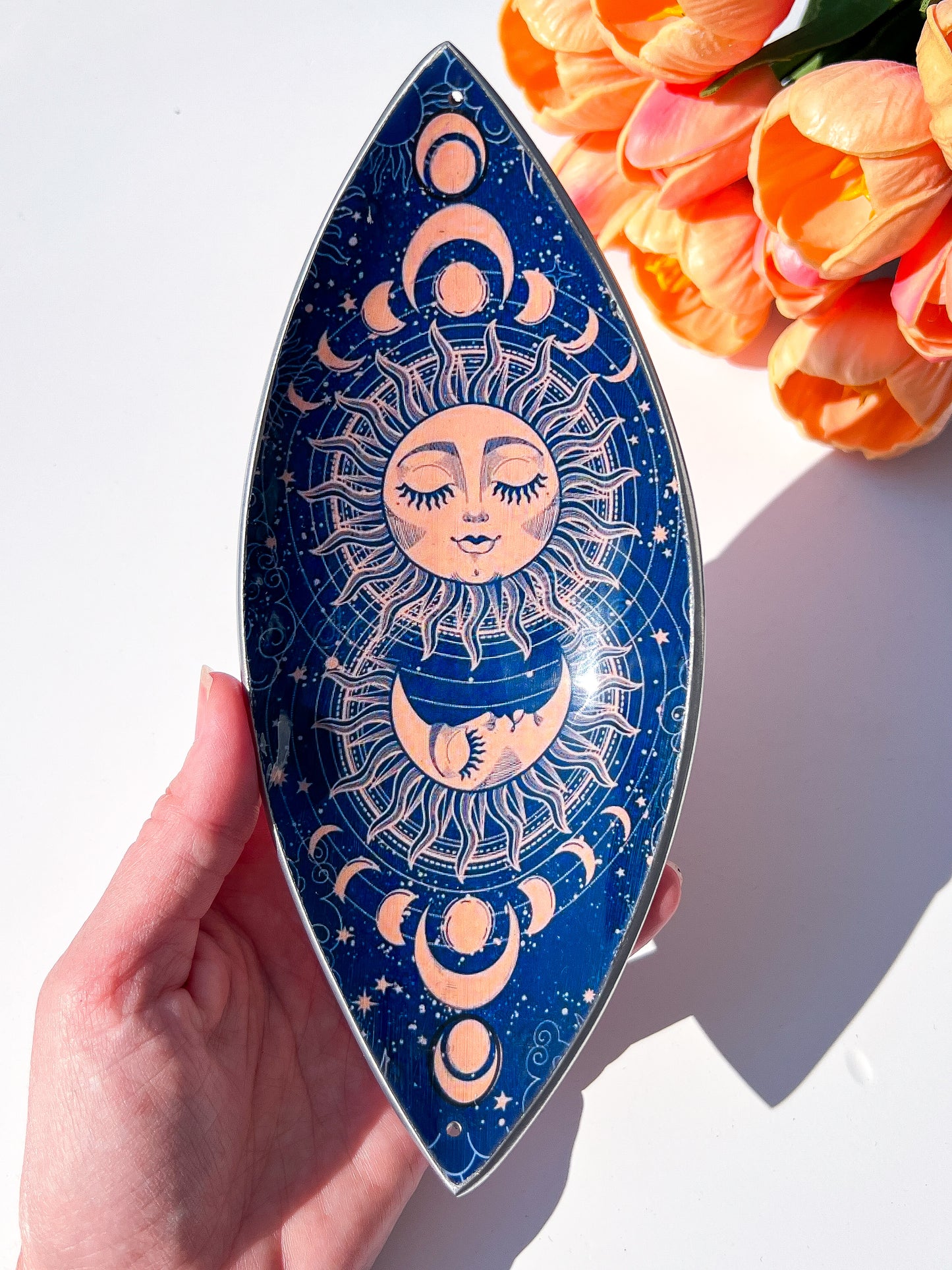 A hand holds a teardrop-shaped decorative plate with a celestial sun and moon design in navy blue and peach, with orange tulips in the top right corner.