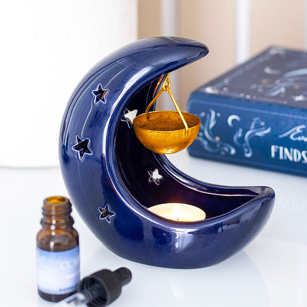 Blue ceramic moon-shaped oil burner with a candle inside, next to a bottle and dropper on a white surface.
