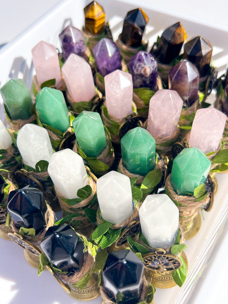 A collection of faceted crystal points in various colors including green, pink, purple, white, black, and amber, each nestled in a small woven basket with green leaves, arranged in a white tray.