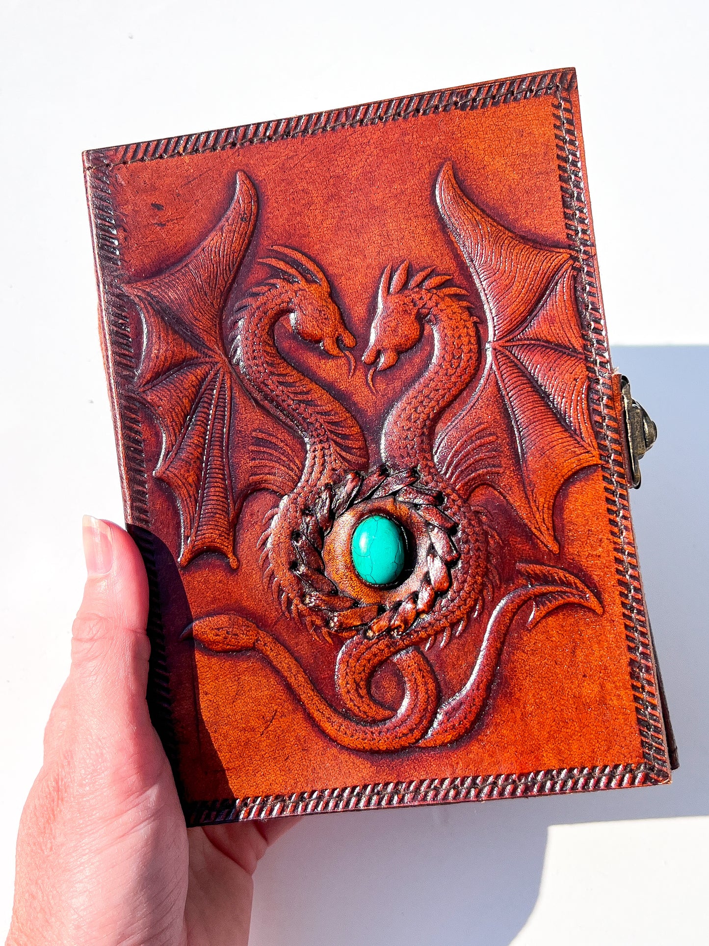 A hand holds a small embossed leather journal featuring two dragons facing each other around a turquoise stone, set against a plain white background.