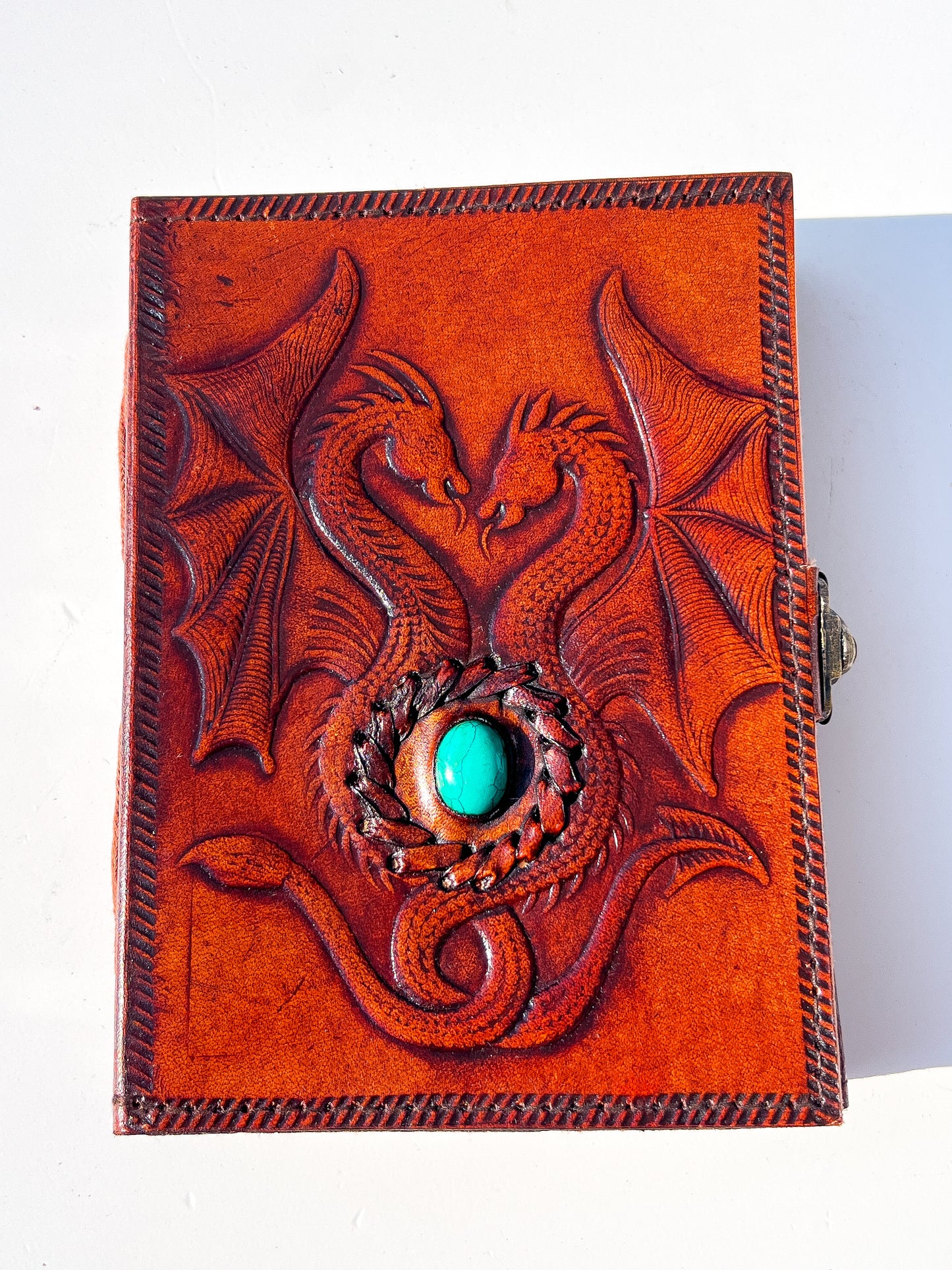 A rectangular brown leather journal featuring embossed dragon designs and a central turquoise stone, photographed on a plain white background.