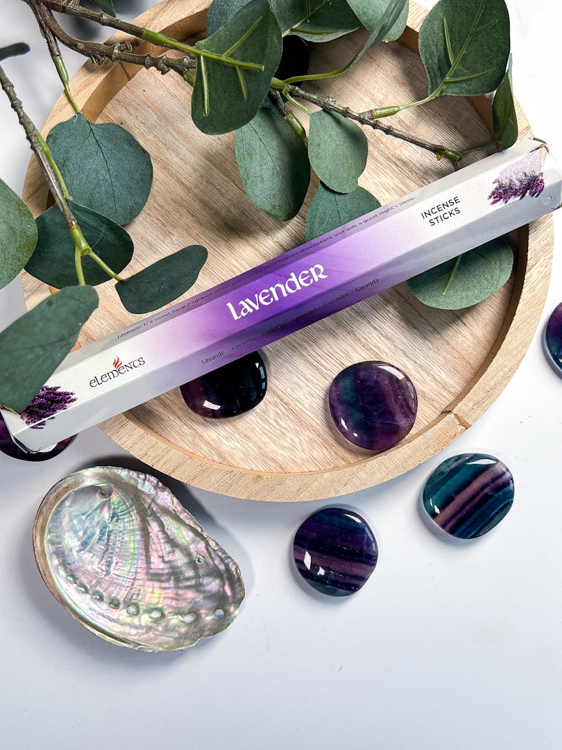 Lavender incense stick with packaging on a wooden coaster with green leaves and decorative stones.