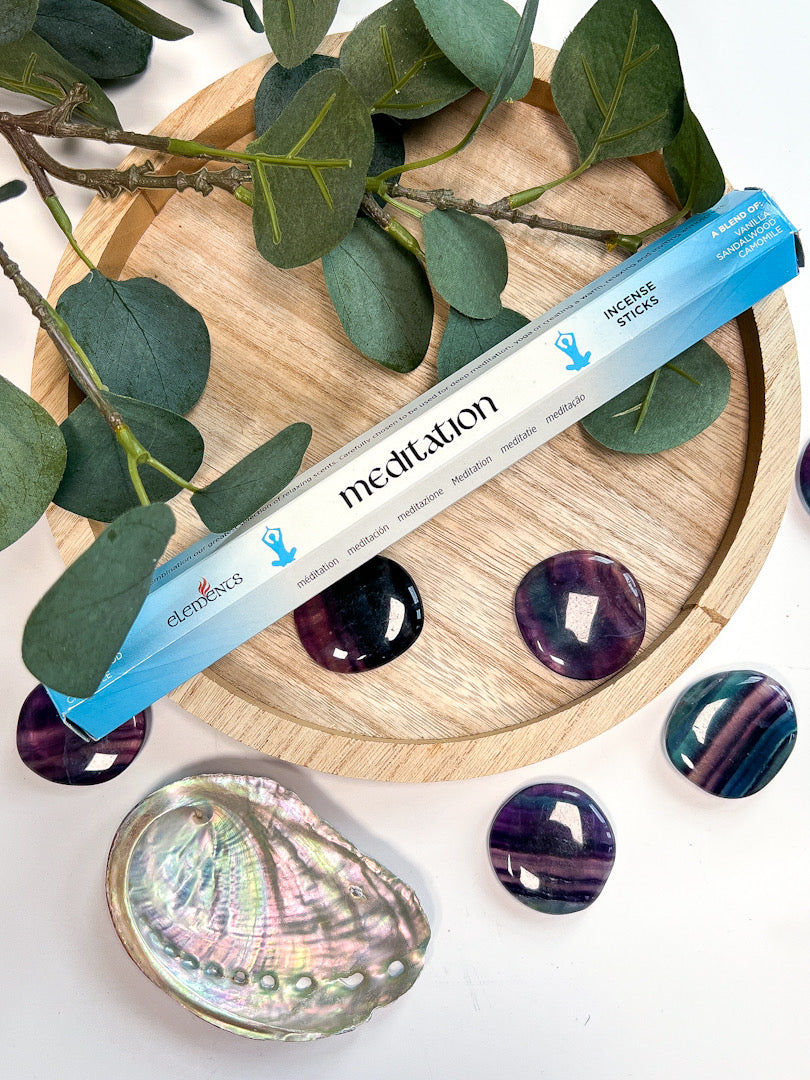 Incense stick labeled 'Meditation' on a wooden tray with green leaves and stones.