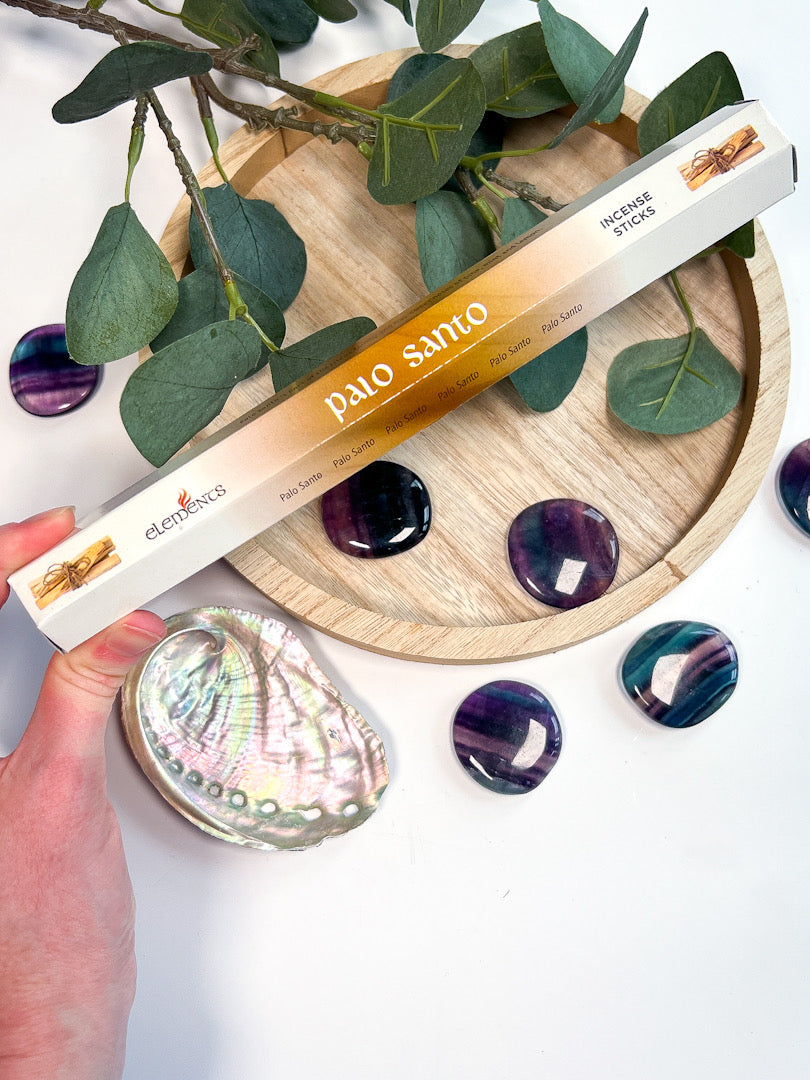 Palo Santo abalone shell with a hand holding it, surrounded by purple stones and green leaves on a wooden board.