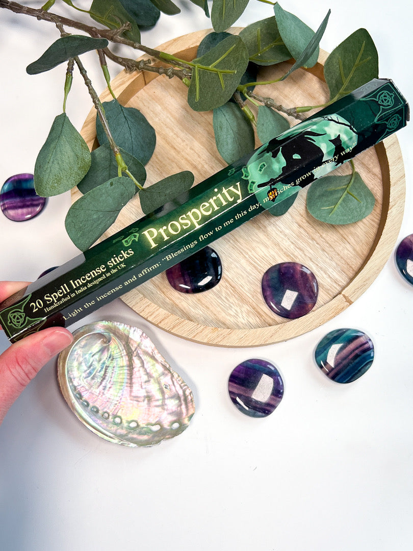 Prosperry incense stick packaging with green design on a wooden coaster with eucalyptus leaves and amethyst stones.