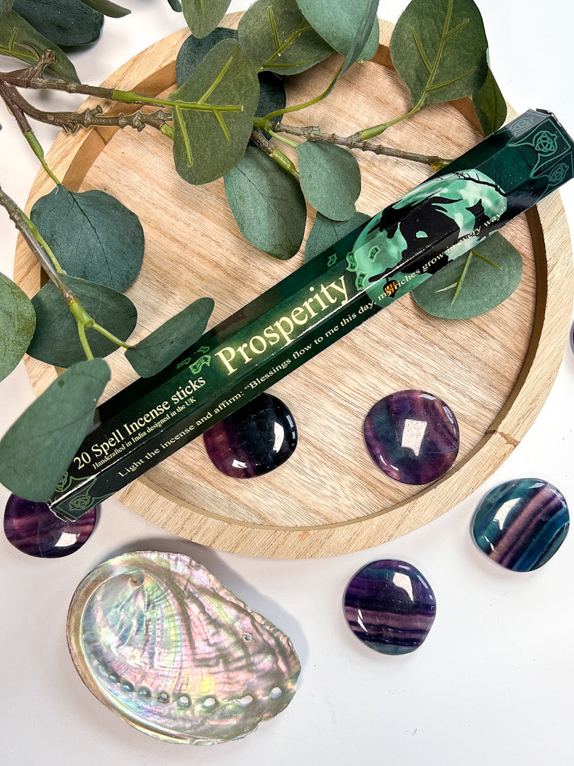 Incense stick labeled 'Prosperity' on a wooden coaster with greenery and stones.