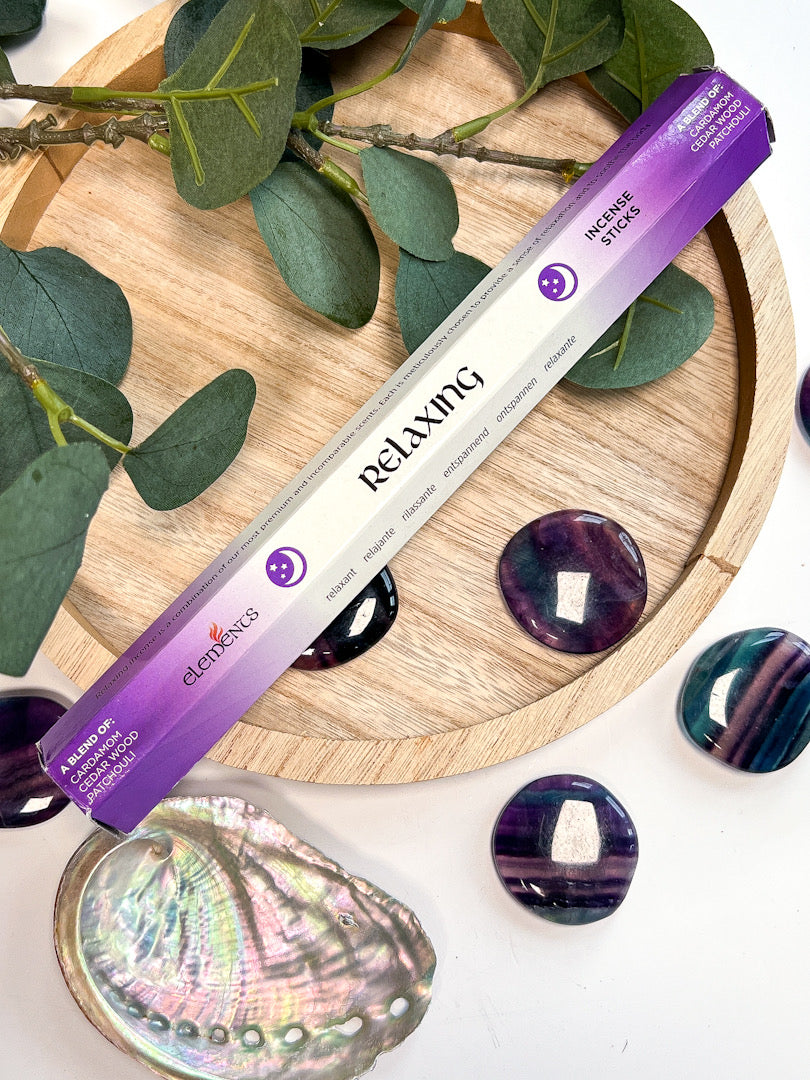 Incense stick labeled 'Relaxing' on a wooden tray with green leaves and purple stones.