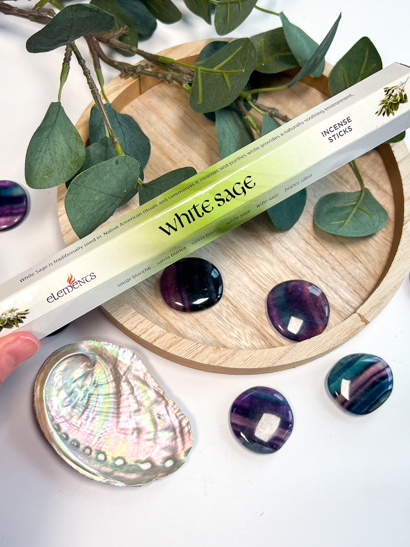 White sage incense sticks with a wooden coaster, green leaves, and purple stones on a white background