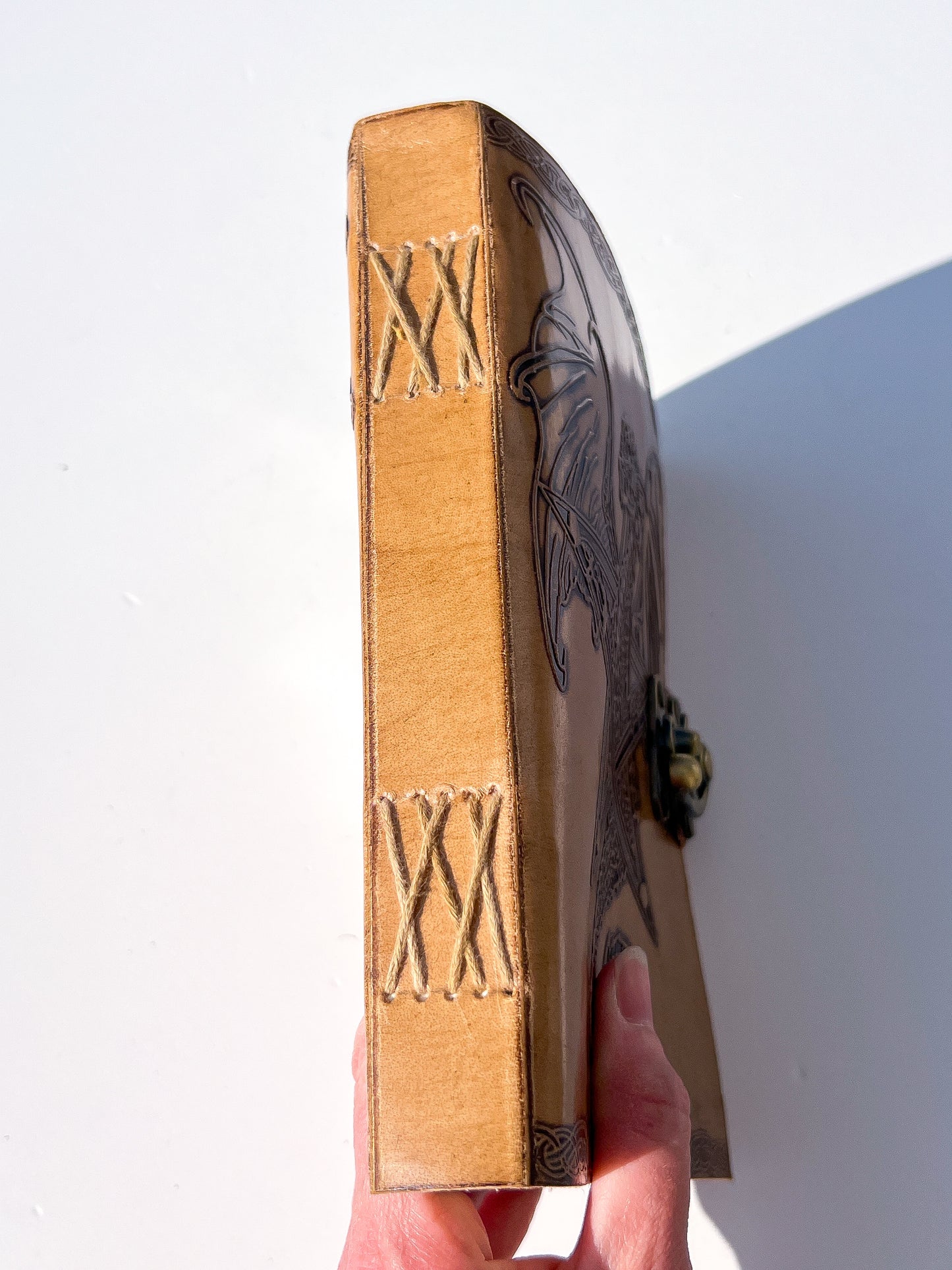A hand holding a leather-bound book or journal upright against a plain white background. The spine is tan leather with decorative stitching and Roman numerals, and the cover has dark engraved artwork and a small metal clasp.