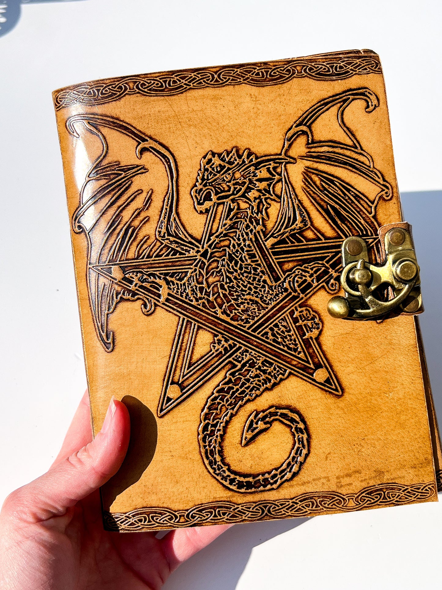 A hand holds a tan leather journal featuring an embossed dark brown dragon over a pentagram design, secured with a brass clasp on a white background.
