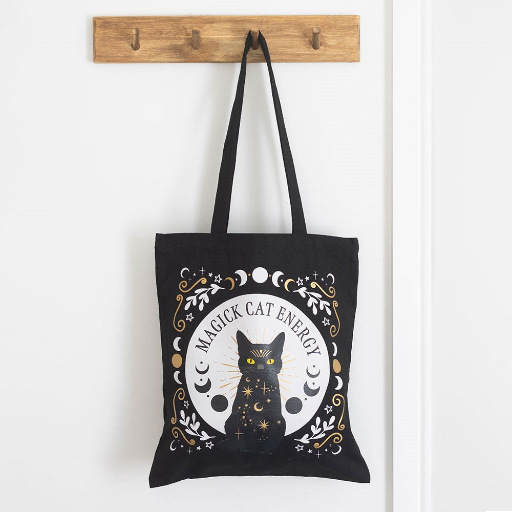 Black tote bag with a cat design and 'Magick Cat Energy' text, hanging on a wooden hook.