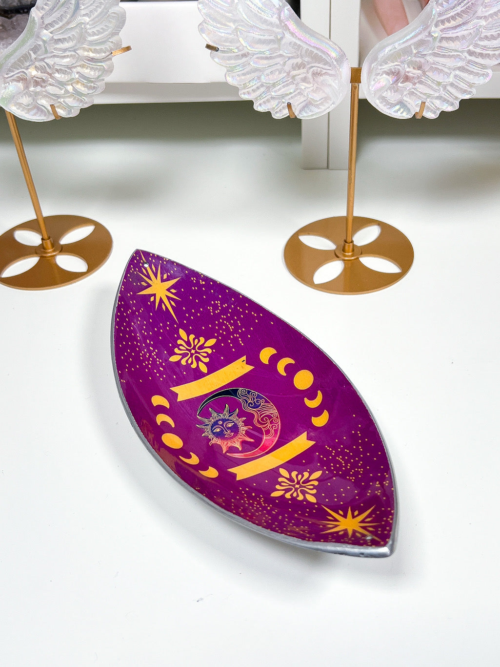 purple sun and moon phases incense stick holder on a white table with angel wings in the background.
