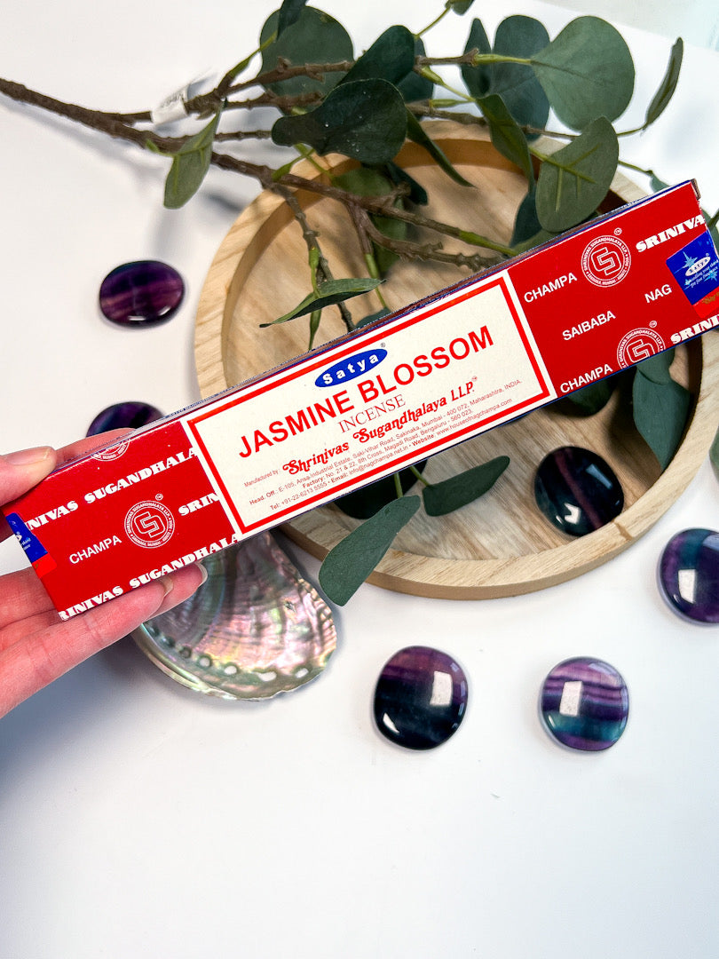 Jasmine Blossom incense stick box held by a hand with decorative stones and greenery in the background.