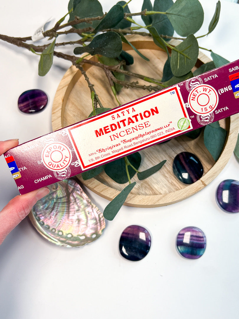 Satya Meditation incense packaging held by a hand with decorative stones and leaves in the background