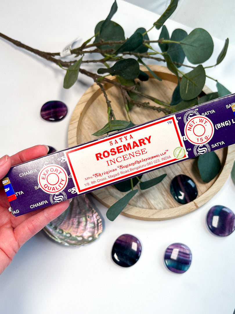 Satya Rosemary incense in hand with wooden tray and greenery behind.