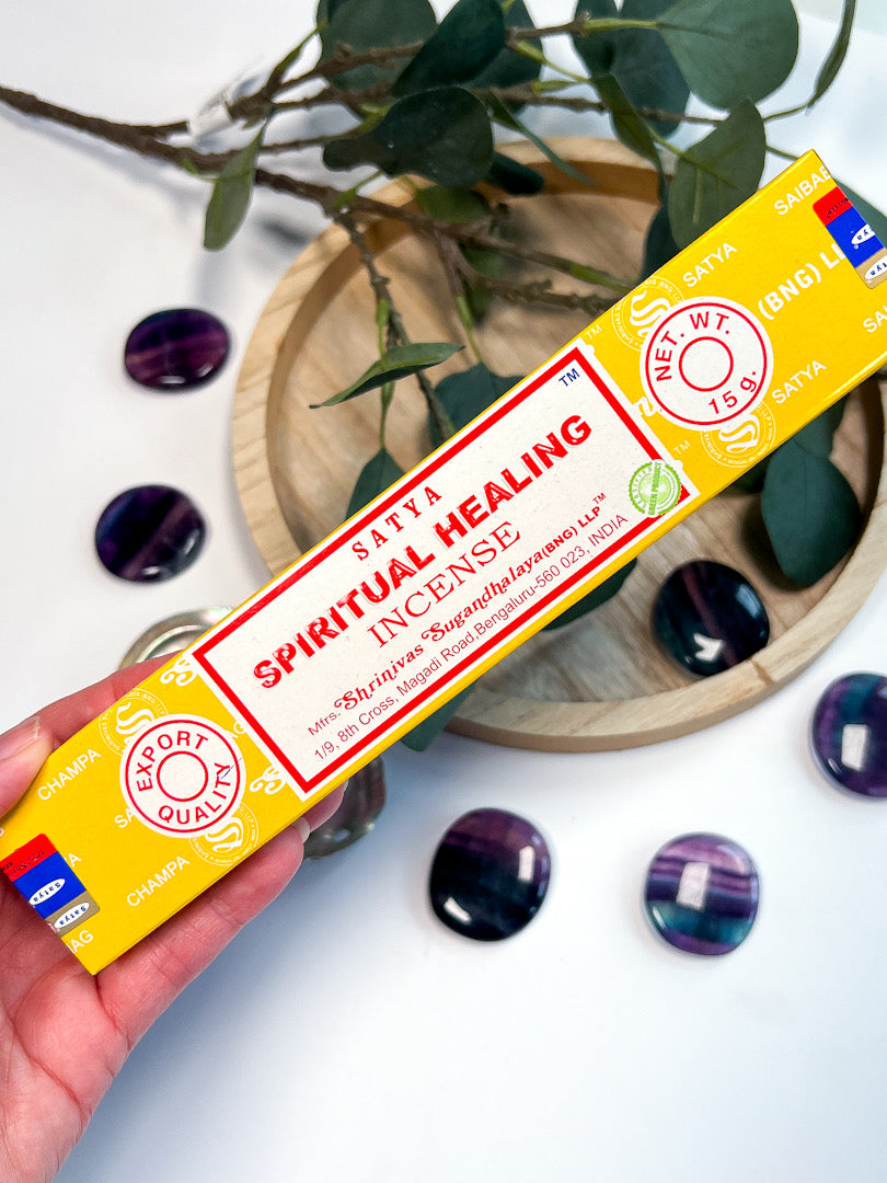 Box of Satya Spiritual Healing Incense held by a hand with decorative stones and plants in the background.