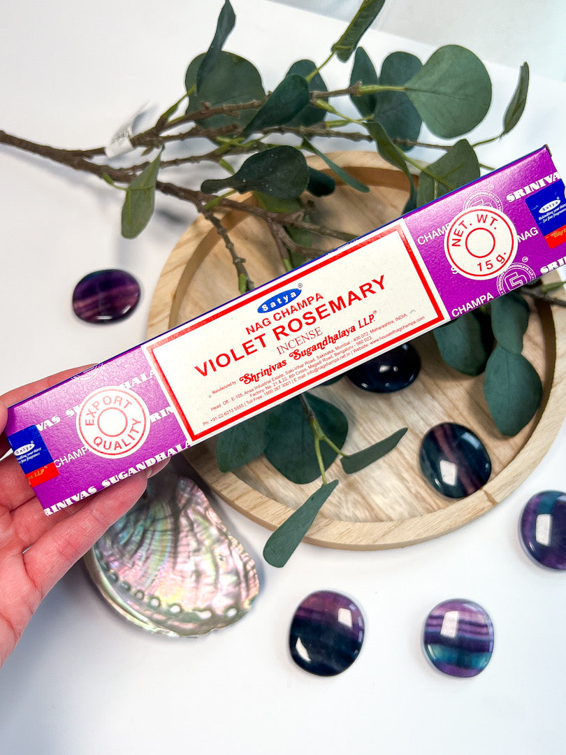 Hand holding a package of Violet Rosemary incense sticks with a decorative background.